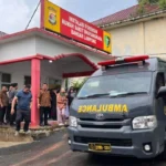 Crowd of people outside the Ruang Forensik at Bhayangkara Hospital Bandar Lampung as a gray ambulance with red emergency lights arrives.