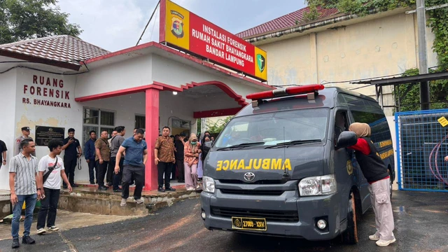 Crowd of people outside the Ruang Forensik at Bhayangkara Hospital Bandar Lampung as a gray ambulance with red emergency lights arrives.