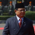 Man in a dark suit and red tie walks on a red carpet at a formal ceremony, wearing a black cap with guards in the background.