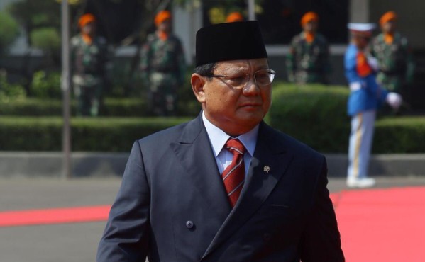 Man in a dark suit and red tie walks on a red carpet at a formal ceremony, wearing a black cap with guards in the background.