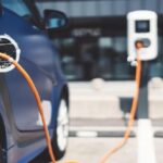 Blue electric car charging at a station with an orange charging cable plugged into the vehicle in a parking area.
