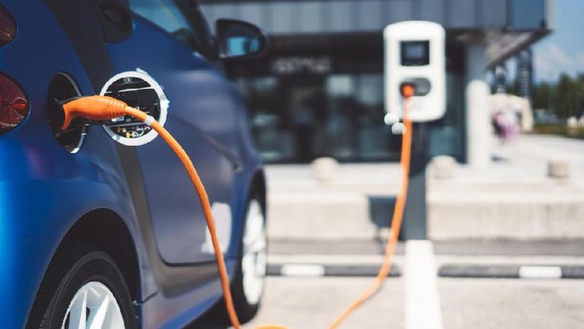 Blue electric car charging at a station with an orange charging cable plugged into the vehicle in a parking area.