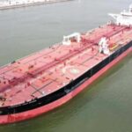 Large red-and-black tanker ship viewed from above sailing in a calm waterway, seen from bow to stern.