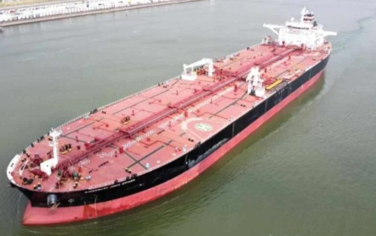 Large red-and-black tanker ship viewed from above sailing in a calm waterway, seen from bow to stern.