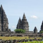 Stone temple complex with tall, stepped spires rising above a low stone wall under a blue sky (Borobudur-like temple).