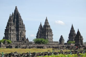 Stone temple complex with tall, stepped spires rising above a low stone wall under a blue sky (Borobudur-like temple).