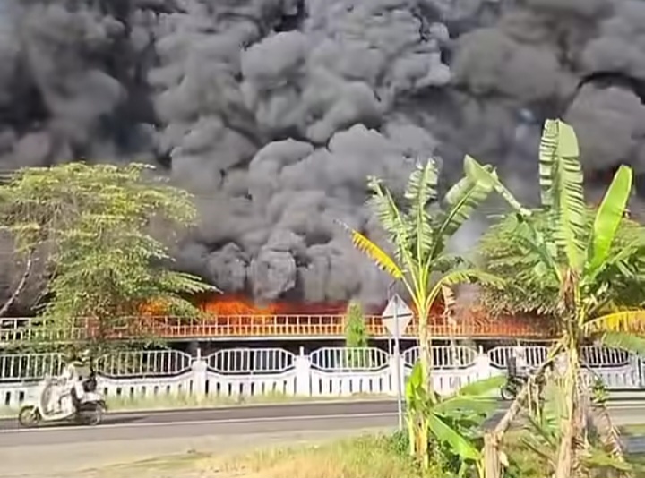 Fire on a building or area behind a white fence, with dense black smoke filling the sky; banana trees in the foreground.