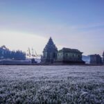 Ancient stone temple ruins in a frost-covered field at dawn, with distant trees and a pale purple sky.