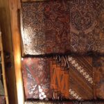Display of batik fabric rolls in warm earth tones with intricate floral and geometric patterns on a wall display.
