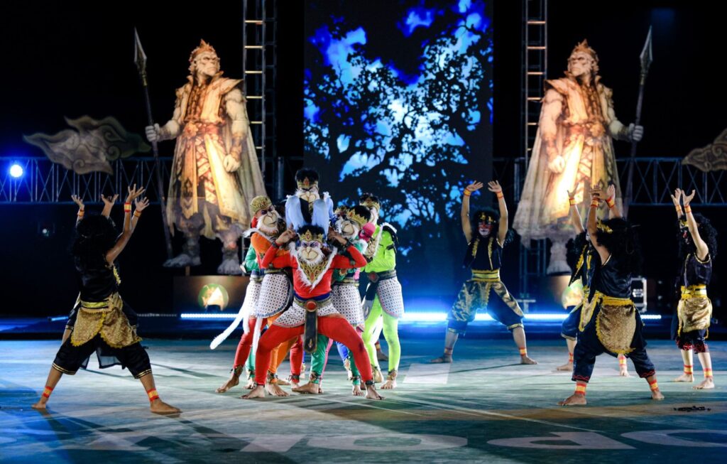 Group of performers in colorful costumes and masks on a stage, with two large statue-like figures and a blue abstract backdrop behind them.