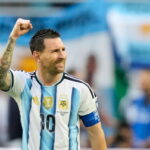 Argentine soccer player in a blue-and-white striped jersey raises a clenched fist in celebration on the field, wearing a captain's armband.