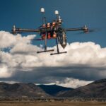Drone in flight over a distant mountain valley with a clear blue sky and scattered clouds behind it.
