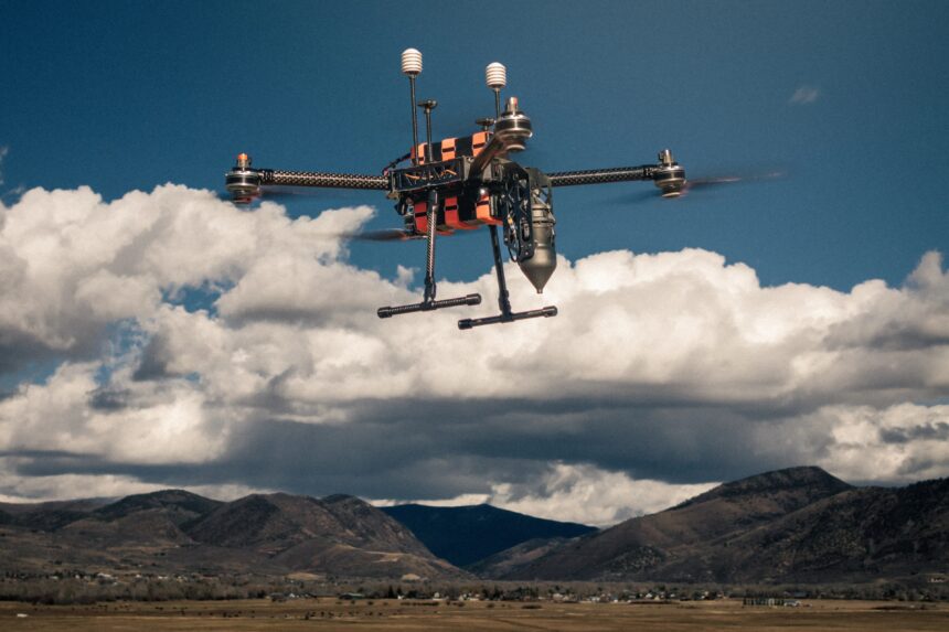 Drone in flight over a distant mountain valley with a clear blue sky and scattered clouds behind it.