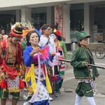 Group of costumed performers in bright outfits and face paint marching down a city street.