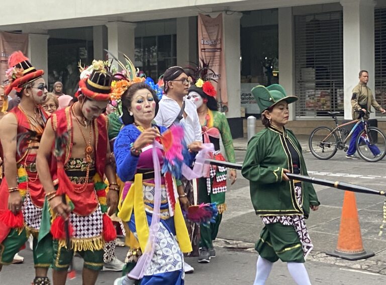 Group of costumed performers in bright outfits and face paint marching down a city street.