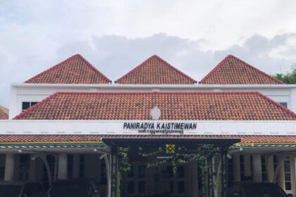 Front view of a low, white building with multiple red-tiled gabled roofs and a covered entrance with a sign above the doors.
