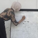 Woman in a light hijab writes on a whiteboard filled with handwritten notes in a classroom setting.