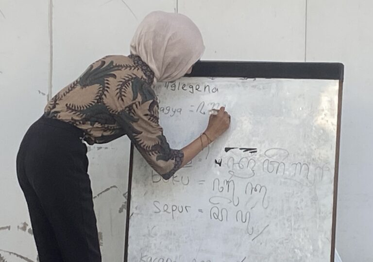 Woman in a light hijab writes on a whiteboard filled with handwritten notes in a classroom setting.