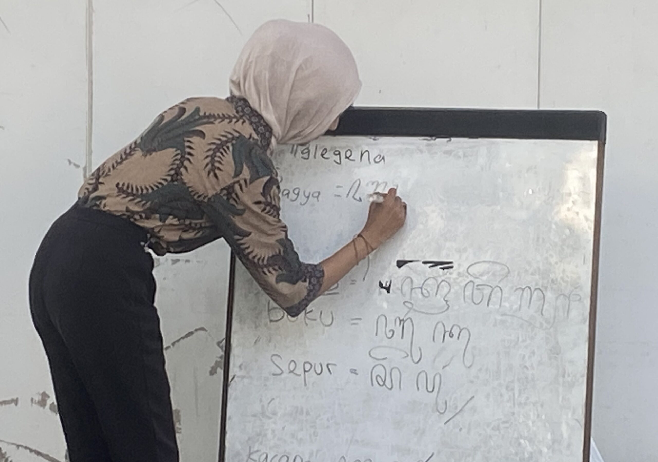 Woman in a light hijab writes on a whiteboard filled with handwritten notes in a classroom setting.