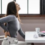 Woman sitting at a desk clutching her lower back in visible discomfort at an office with a laptop and papers nearby