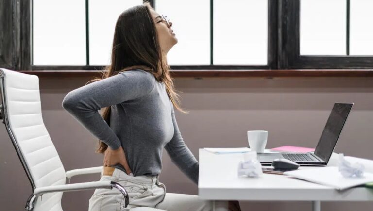 Woman sitting at a desk clutching her lower back in visible discomfort at an office with a laptop and papers nearby