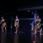 Three dancers in gold-brown traditional costumes perform on a dimly lit stage.