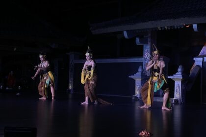 Three dancers in gold-brown traditional costumes perform on a dimly lit stage.