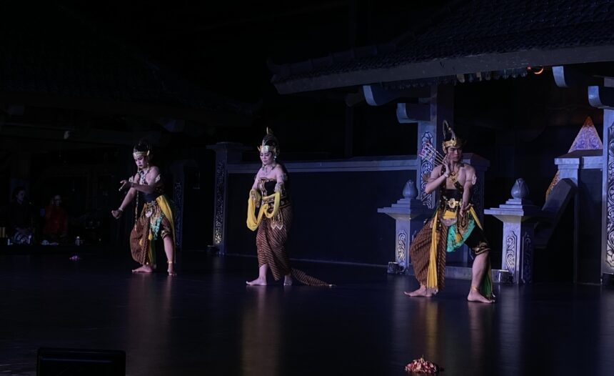 Three dancers in gold-brown traditional costumes perform on a dimly lit stage.