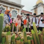 Group of people in traditional clothing gathered around a display of cut bamboo stalks and coconuts during a cultural ceremony outside a mosque-like building.
