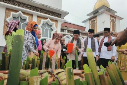 Group of people in traditional clothing gathered around a display of cut bamboo stalks and coconuts during a cultural ceremony outside a mosque-like building.