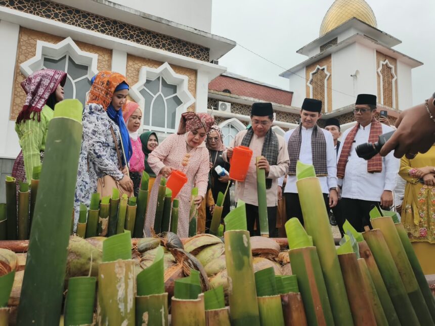 Group of people in traditional clothing gathered around a display of cut bamboo stalks and coconuts during a cultural ceremony outside a mosque-like building.