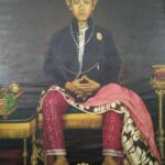 Portrait of a seated person in traditional attire: green fez, dark jacket with pearls, pink patterned pants, and a decorative shawl draped over the lap; hands folded.