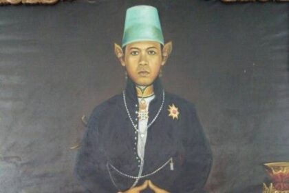Portrait of a seated person in traditional attire: green fez, dark jacket with pearls, pink patterned pants, and a decorative shawl draped over the lap; hands folded.