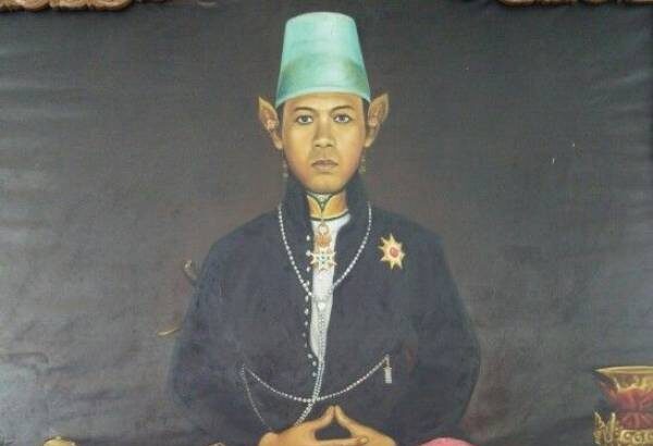 Portrait of a seated person in traditional attire: green fez, dark jacket with pearls, pink patterned pants, and a decorative shawl draped over the lap; hands folded.
