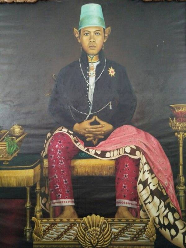 Portrait of a seated person in traditional attire: green fez, dark jacket with pearls, pink patterned pants, and a decorative shawl draped over the lap; hands folded.