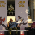 Three panelists seated on stage during Festival Literasi Jogja 2026, with a man speaking into a microphone between a woman in a hijab and a man in a pink shirt, backdrop shows 'Festival Literasi' and 'JOGJA 2026' banners.