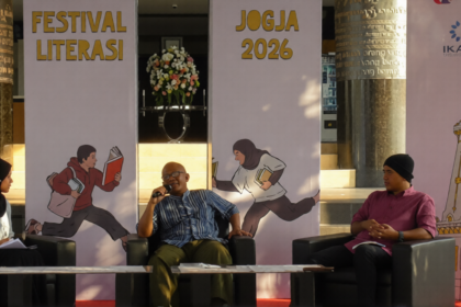 Three panelists seated on stage during Festival Literasi Jogja 2026, with a man speaking into a microphone between a woman in a hijab and a man in a pink shirt, backdrop shows 'Festival Literasi' and 'JOGJA 2026' banners.