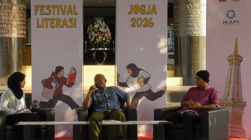 Three panelists seated on stage during Festival Literasi Jogja 2026, with a man speaking into a microphone between a woman in a hijab and a man in a pink shirt, backdrop shows 'Festival Literasi' and 'JOGJA 2026' banners.