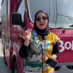 Woman in a black hijab and sunglasses stands beside a pink truck with white lettering, holding a smartphone and gesturing with her finger.