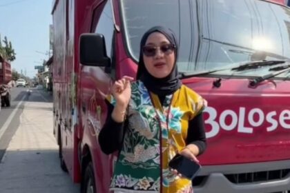 Woman in a black hijab and sunglasses stands beside a pink truck with white lettering, holding a smartphone and gesturing with her finger.