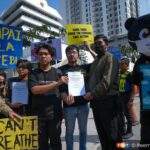 Group of protesters holding bright yellow placards; two men exchange a document while a panda mascot stands on the right with a sign about haze