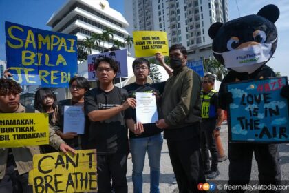 Group of protesters holding bright yellow placards; two men exchange a document while a panda mascot stands on the right with a sign about haze