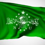 Green flag with a white globe emblem, rope knot, stars, and Arabic script across the center, flapping in wind.