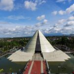 Pyramid-shaped white temple rising from a platform, surrounded by a rectangular moat and a red-carpeted walkway leading to stairs under a blue sky.
