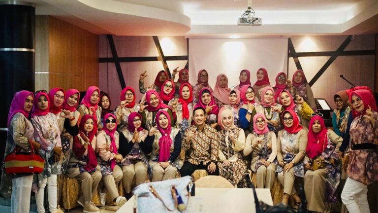 Group of women wearing pink hijabs and colorful outfits posing for a group photo at an indoor event. A few men sit in front center.