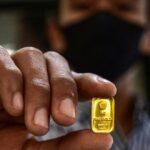 Close-up of a person in a mask holding a small gold bullion bar between their fingers.
