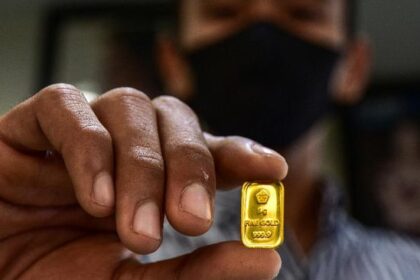 Close-up of a person in a mask holding a small gold bullion bar between their fingers.