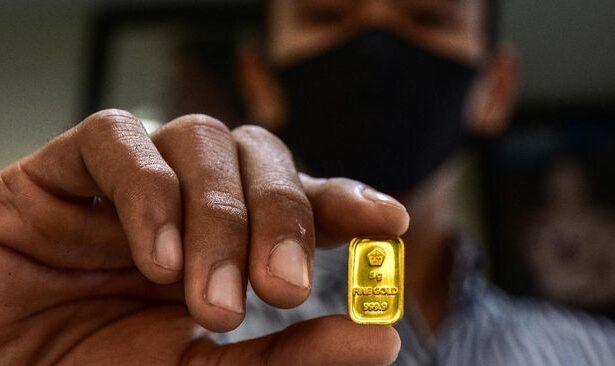 Close-up of a person in a mask holding a small gold bullion bar between their fingers.