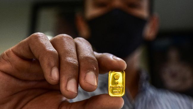 Close-up of a person in a mask holding a small gold bullion bar between their fingers.