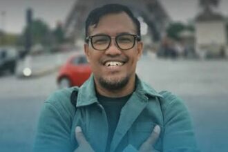 Friendly portrait of a man with glasses wearing a teal jacket, arms crossed, outdoors with a blurred city street behind him.